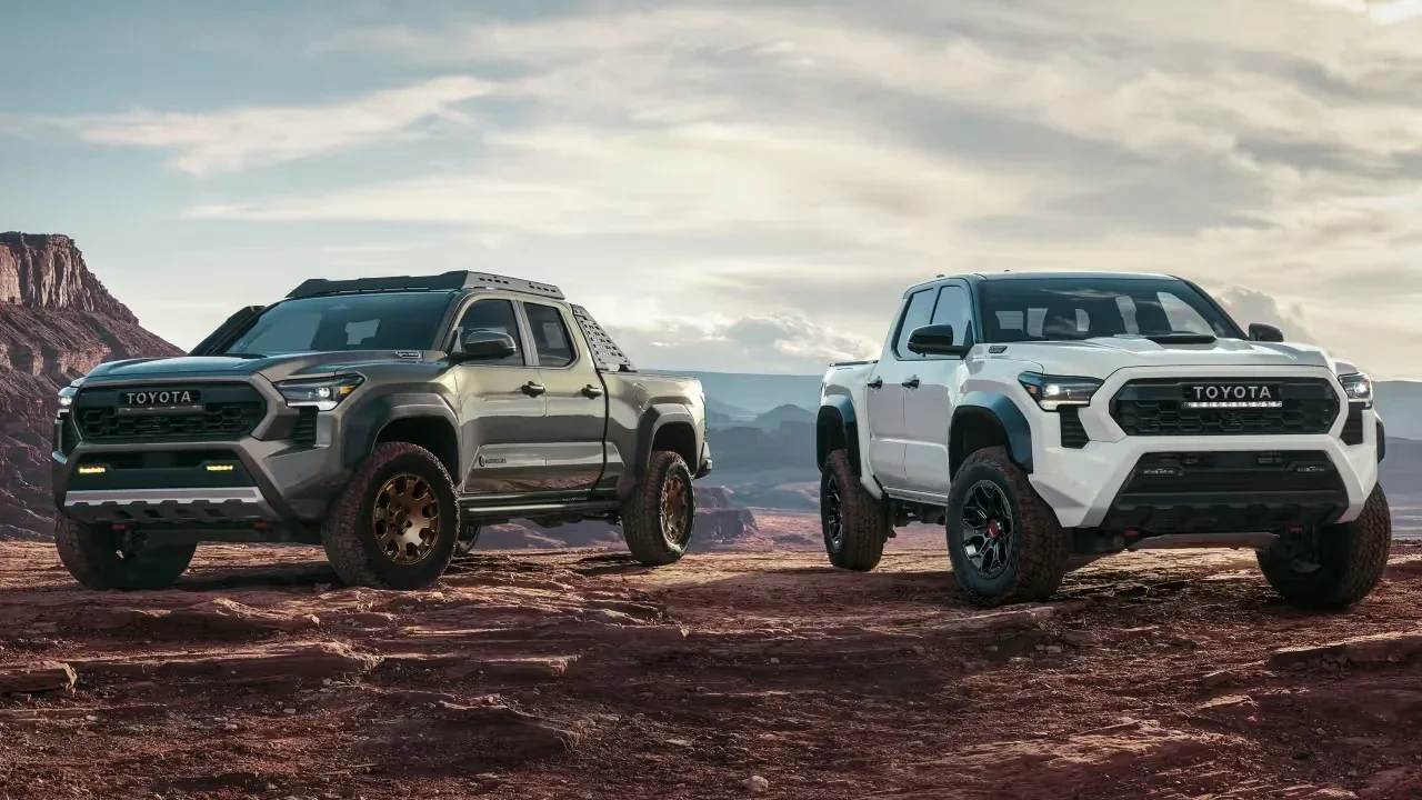 Two 2025 Toyota Tacoma i-Force MAX trucks parked on rugged desert terrain at sunset—one in metallic gray, the other in white—highlighting their aggressive off-road stance, bold front grilles, and hybrid performance design.